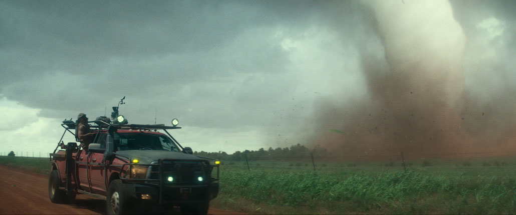 A vertical shot from the movie 'Twisters' shows a red truck on the right equipped with tornado tracking and additional lights. A man leans halfway out the window to look toward a nearby tornado on the right side of the image.